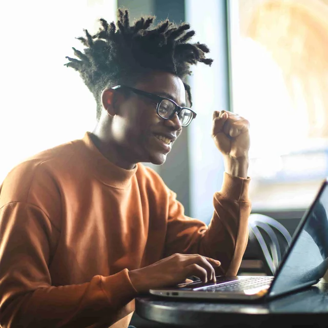 Young man smiling and celebrating while taking an online language speaking test on his laptop, symbolizing success through Berlitz assessment tools.